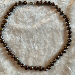 May include: A necklace featuring dark brown, irregularly shaped pearls strung on a dark cord. The necklace is secured with a silver-colored clasp. The pearls have a lustrous sheen, and the necklace is displayed against a light-colored, textured background.