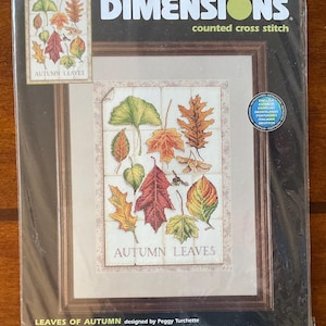 May include: A Dimensions counted cross stitch kit titled Autumn Leaves featuring a variety of colorful fall foliage on a white grid background. The package includes cotton thread, 14 count ivory Aida, a needle, and instructions for a 10 by 14 inch design.