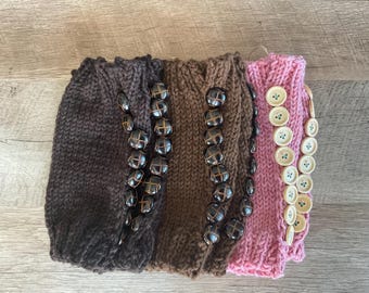 May include: Three knitted boot cuffs arranged side by side. The first is dark brown, the second is medium brown, and the third is light pink. Each cuff features a vertical row of round buttons attached along the outer edge.