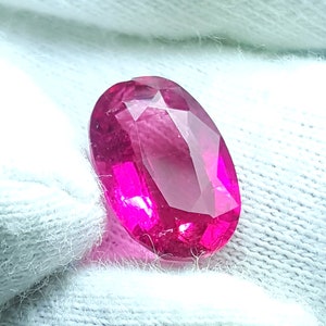 May include: A vibrant, oval-cut gemstone with a deep, saturated magenta-pink hue. The stone features a faceted surface that reflects light, creating internal brilliance and clarity. It is held against a dark background, showcasing its colour.