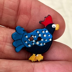 May include: A colorful, decorative button in the shape of a chicken. The button features a red comb, yellow beak and feet, and a blue body with white polka dots. The button has two holes for sewing.