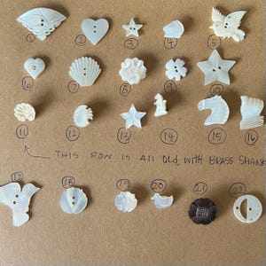 May include: A collection of 22 vintage white mother of pearl buttons with various shapes and designs, including a star, a heart, a bird, a shell, a flower, a fish, a horse, and a duck. The buttons are arranged in rows on a brown surface. The text "THIS ROW IS ALL OLD, WITH BRASS SHANK" is written below the buttons.