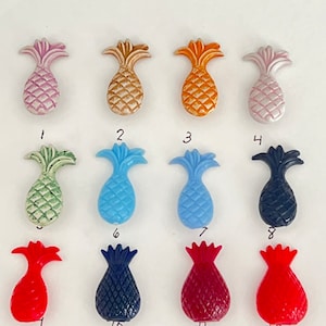 May include: Twelve pineapple-shaped buttons in various colors, including pink, orange, green, blue, red, and navy blue. The buttons are arranged in three rows of four.