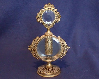 May include: An antique perfume bottle with a clear glass body and ornate gold-colored filigree metalwork. The bottle has a round base, a central oval section, and a smaller, diamond-shaped top, all framed by decorative metal. The design is symmetrical and elegant.