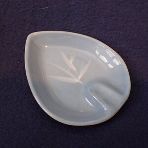May include: A light blue, leaf-shaped ashtray with a white floral design. The ashtray has a curved indentation for resting cigarettes. The ceramic ashtray is a vintage piece.