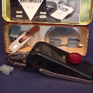 May include: A black buttonhole sewing machine with a red button and a silver lever. The machine is in a tin box with a label that says "Buttonholer B-3". The box also contains various sewing attachments.