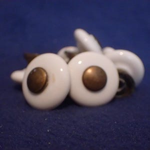 May include: A collection of white ceramic cabinet knobs with brass-colored centers. The knobs are round and have a classic design. The hardware is set against a dark blue background.