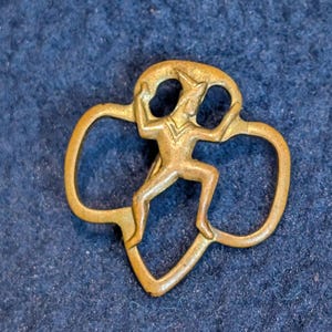 May include: A vintage brass-toned pin featuring a stylized figure within a trefoil design. The figure is depicted in a dynamic pose, with arms raised. The pin has a slightly aged appearance, suggesting it is an antique or vintage item.