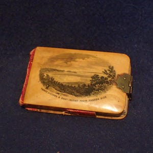 May include: A small, wooden photo album with a hinged clasp. The cover features a black and white illustration of a landscape with the text "Three Sisters & Goat Island from Canada Side".