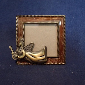 May include: Square photo frame with a bronze-colored angel playing a stringed instrument. The frame has a brown border with a dotted pattern and a bronze-colored angel attached to the frame.