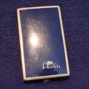 May include: A vintage Avon compact with a gold trim and a blue enamel finish. The compact has a small house design and the word "Avon" in gold lettering.