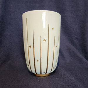 May include: A white ceramic vase with vertical gold stripes and star accents. The vase has a gold base and a cylindrical shape. The design is symmetrical, with the gold elements evenly spaced around the vase.