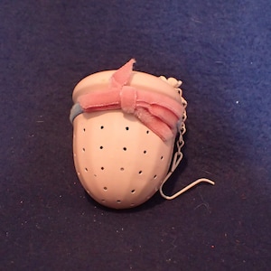 May include: A white tea infuser with small holes, a pink bow, and a chain. The infuser has a rounded shape and is designed for steeping loose leaf tea. The bow adds a decorative touch to the tea accessory.