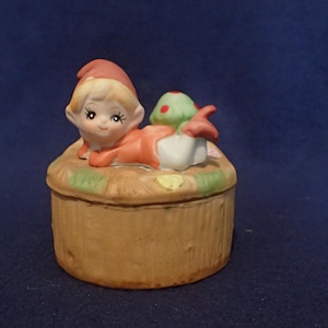 May include: A ceramic trinket box with a small elf figurine lying on top. The elf is wearing a red hat and a red and white outfit. The elf is holding a green and red mushroom. The box is brown and has a textured surface.