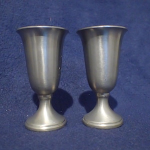 May include: Two vintage pewter goblets with a flared rim and a pedestal base. The goblets have a smooth, metallic finish and are displayed against a blue background. The stemware is likely used for serving beverages.