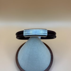 May include: A dark brown leather bracelet with a silver-colored clasp. The clasp has a textured design. The bracelet is displayed on a light blue, cone-shaped stand with a dark brown wooden base.