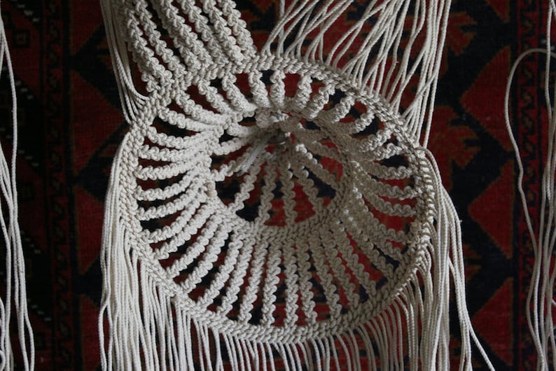 K&ouml;nnte beinhalten: Ein wei&szlig;er Makramee-Wandbehang mit einem runden Design und Fransen. Der Makramee ist aus dickem Baumwollgarn gefertigt und hat ein zartes, filigranes Muster.