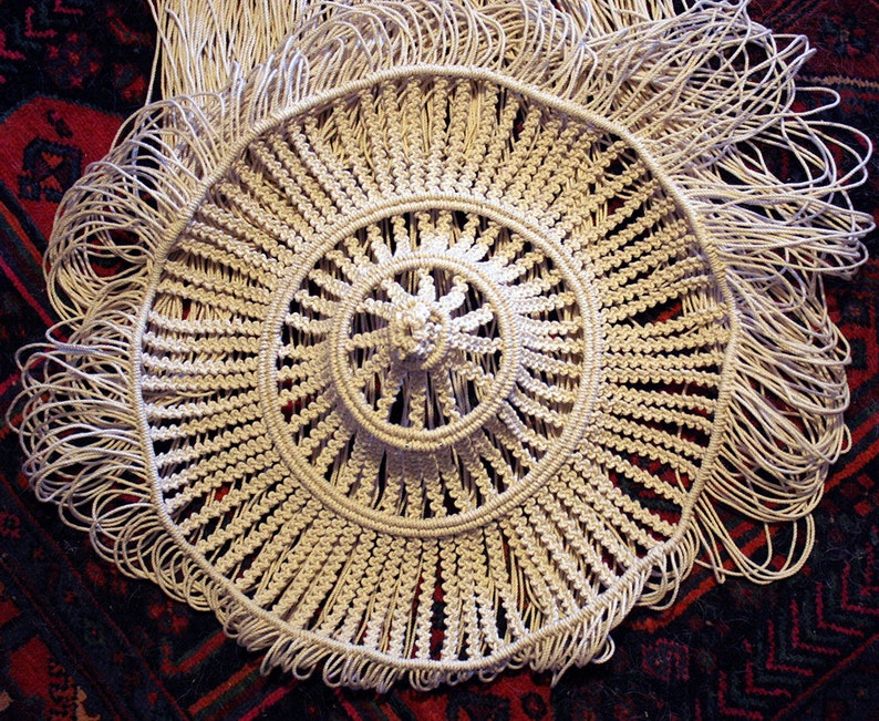 K&ouml;nnte beinhalten: Ein wei&szlig;es geh&auml;keltes Mandala mit einem Fransenrand. Das Mandala besteht aus einer Reihe konzentrischer Kreise mit einem Mittelpunkt.