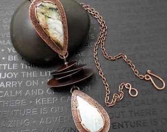 May include: A copper necklace with a layered pendant featuring a green and white agate stone and a teardrop-shaped pendant with a mirrored surface. The necklace is on a dark gray background.