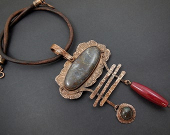 May include: A copper necklace with a large, oval-shaped agate stone pendant. The pendant is surrounded by a textured copper frame and hangs from a leather cord. The necklace also features a smaller copper pendant with a green stone and a long, red agate bead.