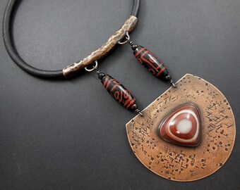 May include: A copper necklace with a large, hammered, half-circle pendant featuring a red and white agate stone. The pendant is attached to a black leather cord with a hammered copper bar and two black and red agate beads.