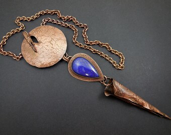 May include: A copper necklace with a large round pendant, a teardrop pendant with a blue lapis lazuli stone, and a long, pointed copper pendant. The necklace is on a chain.