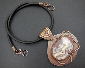 May include: A copper necklace with a large, oval-shaped pendant featuring a colorful agate stone set in a copper frame. The pendant is attached to a black leather cord.