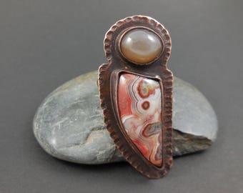 May include: A copper pendant with a red, white, and brown agate stone set in a copper frame. The pendant is shaped like a teardrop and has a textured surface.