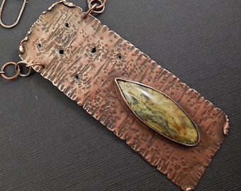 May include: A copper pendant with a textured surface and a green and brown stone set in the center. The pendant is attached to a copper chain.
