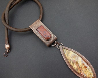 May include: A leather cord necklace with a copper pendant featuring two stones. The top stone is a reddish-brown color and the bottom stone is a light brown with darker brown and yellow markings.