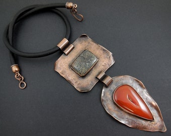 May include: A copper necklace with a black leather cord and a pendant featuring two irregularly shaped stones set in hammered copper. The larger stone is a dark green and the smaller stone is a reddish-orange.