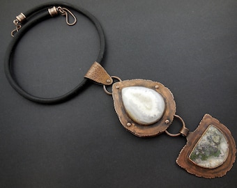 May include: A copper necklace with a pendant featuring two agate stones. The larger stone is white with gray veins, and the smaller stone is green with white veins. The necklace is made of black leather cord.
