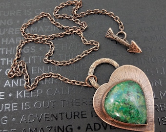 May include: A copper heart-shaped pendant necklace with a green gemstone set in the center. The necklace features a copper chain and a small arrow charm.