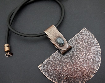 May include: A copper necklace with a hammered, half-circle pendant and a dark gray gemstone. The necklace is on a black leather cord.