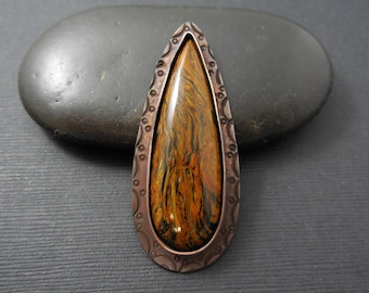 May include: A teardrop-shaped pendant with a copper frame and a tiger's eye stone. The frame has a decorative design with small circles and swirls.