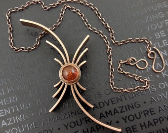 May include: A copper necklace with a pendant featuring a geometric design with a central amber stone. The necklace is on a dark gray surface with text that reads "You're amazing. Adventure is out there. Keep calm and carry on. Be happy. Be things. The little things. The joy of things."