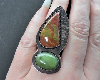 May include: A silver ring with two stones. The larger stone is a teardrop shape with red, green, and brown colors. The smaller stone is round and green. The ring is on a finger.