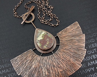 May include: A copper necklace with a large, fan-shaped pendant and a teardrop-shaped stone with green and brown markings. The chain is made of small, linked copper circles.