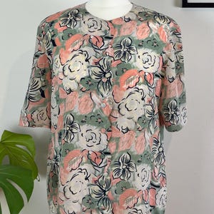 May include: A short-sleeved blouse featuring a scalloped button-down front closure and a round neckline. The fabric displays a repeating floral pattern in shades of sage green, soft pink, and black outlines on a light, textured off-white background.