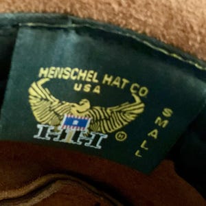 May include: A close-up of a dark green tag from Henschel Hat Co. USA. The tag features a gold eagle emblem with a blue and white flag, and the text "HHH". The size "SMALL" is also visible.