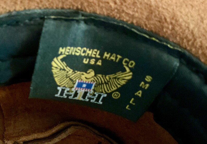 May include: A close-up of a dark green tag from Henschel Hat Co. USA. The tag features a gold eagle emblem with a blue and white flag, and the text "HHH". The size "SMALL" is also visible.