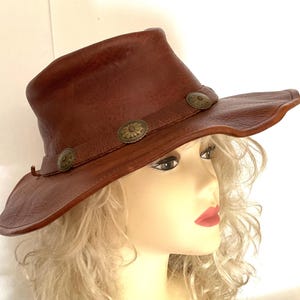 May include: A brown leather hat with a wide brim and a slightly curved crown. The hat features three decorative metal buttons along the brim. The hat is a western-style hat.