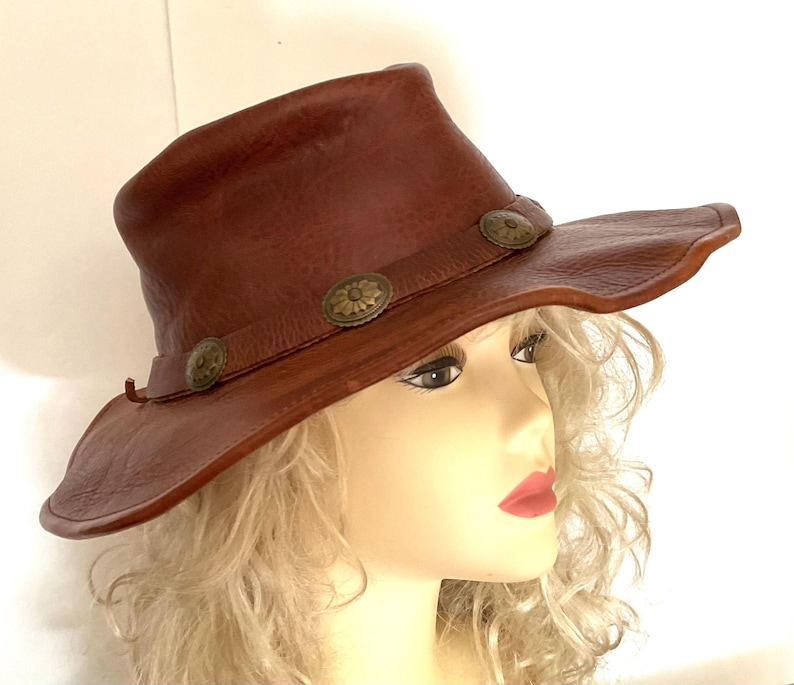 May include: A brown leather hat with a wide brim and a slightly curved crown. The hat features three decorative metal buttons along the brim. The hat is a western-style hat.