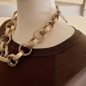 Puede incluir: Un collar dorado y grueso con eslabones texturizados se exhibe en un maniqu&iacute;. El collar es una pieza llamativa, con eslabones grandes e interconectados. El maniqu&iacute; lleva una camiseta marr&oacute;n, creando contraste con el collar dorado.