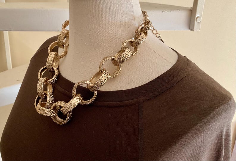 Puede incluir: Un collar dorado y grueso con eslabones texturizados se exhibe en un maniqu&iacute;. El collar es una pieza llamativa, con eslabones grandes e interconectados. El maniqu&iacute; lleva una camiseta marr&oacute;n, creando contraste con el collar dorado.