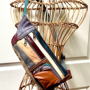 May include: A multi-colored leather fanny pack featuring a patchwork design of purple, burgundy, tan, grey, cream, and teal panels. The bag includes two zippered compartments on the front, a smaller zippered pocket, and an adjustable teal strap.