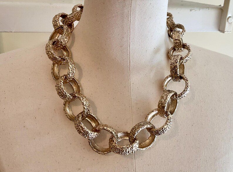 Puede incluir: Un collar grueso en tono dorado con eslabones grandes y texturizados. El collar se exhibe en un maniqu&iacute;, mostrando su dise&ntilde;o audaz. Esta pieza a&ntilde;ade un toque de glamour vintage a cualquier atuendo.