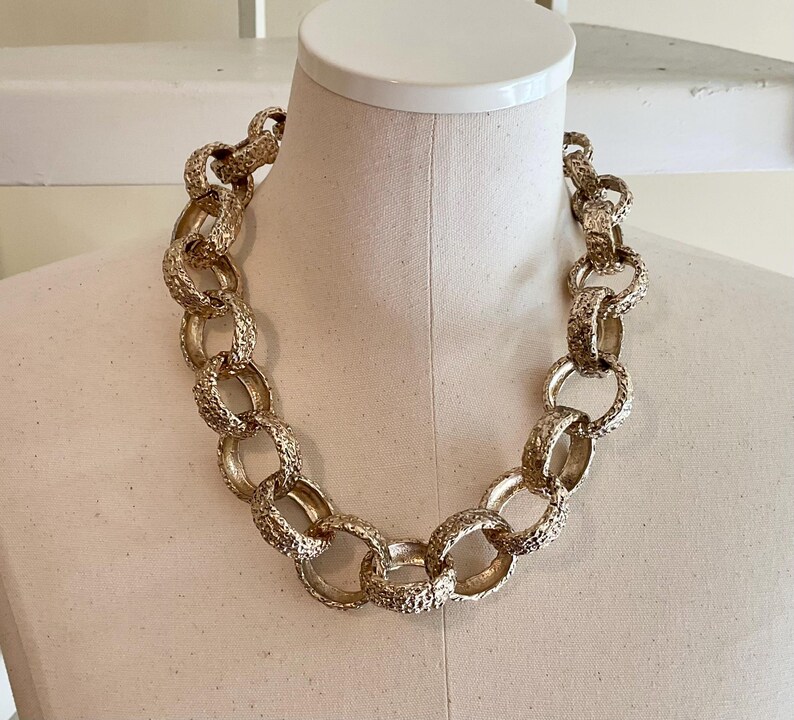 Puede incluir: Un collar dorado con eslabones texturizados grandes. La cadena gruesa se exhibe en un maniqu&iacute;, mostrando su dise&ntilde;o audaz. El collar es una pieza de joyer&iacute;a llamativa.