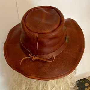 May include: A brown leather hat with a wide brim and a rounded crown. The hat features a decorative metal emblem and a leather tie. The hat's texture is rough and worn, suggesting a vintage or rustic style.