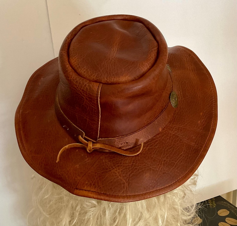 May include: A brown leather hat with a wide brim and a rounded crown. The hat features a decorative metal emblem and a leather tie. The hat's texture is rough and worn, suggesting a vintage or rustic style.
